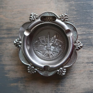 Könnte beinhalten: Silberfarbener Aschenbecher mit einem geprägten Design von Pariser Wahrzeichen, darunter der Eiffelturm. Der Aschenbecher hat einen dekorativen, gewellten Rand und einen kleinen Griff. Er hat drei Zigarettenablagen.