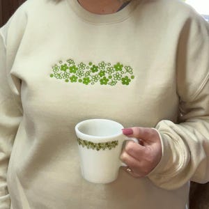 以下が含まれることがあります： 前面に緑色の花の模様がついたベージュのトレーナー。人が緑色の花の模様がついた白いマグカップを持っている。