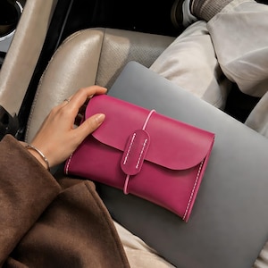 May include: A magenta leather clutch with a flap closure and white stitching. The clutch is secured with a pink elastic band. A gray laptop is beneath the clutch. A person is holding the clutch in a car.