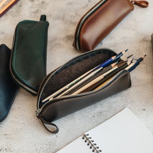 Op de afbeelding: Verschillende leren etuis in diverse kleuren, waaronder bruin, zwart en groen, worden getoond. E&eacute;n etui is open en toont kleurpotloden. Elke etui heeft een ritssluiting en een kleine lus. Een spiraalgebonden notitieboek is ook aanwezig.