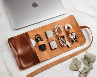 May include: A silver laptop with an Apple logo sits above a tan leather roll-up organizer. The organizer contains a black charging cable, a silver USB-C hub, and two white charging cables, each secured by a small leather strap and metal snap.