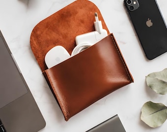 May include: A brown leather pouch with a flap closure containing a white computer mouse and a white power adapter with a coiled cable. The pouch is constructed from smooth, tan-colored leather with visible stitching along the side edges.