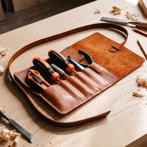 May include: A brown leather tool roll containing various tools, such as screwdrivers, pliers, and a utility knife. The tools have black and orange handles. The roll is open, displaying the tools on a wooden surface.