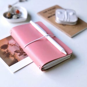 Könnte beinhalten: Ein rosafarbenes Notizbuch mit Ledereinband, das mit einem weißen Gummiband verschlossen ist und auf dem ein weißer Stift liegt. Das Notizbuch liegt neben einem rechteckigen Foto einer Rose und einer kleinen weißen herzförmigen Schale mit Steinen.