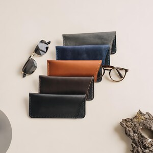 Puede incluir: Una colección de cinco estuches rectangulares de cuero para gafas en tonos gris, azul, tostado, marrón oscuro y negro, cada uno con costuras visibles en los bordes. Dos pares de gafas están colocados cerca de los estuches.