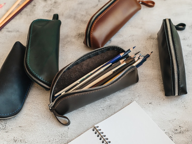Op de afbeelding: Verschillende leren etuis in diverse kleuren, waaronder bruin, zwart en groen, worden getoond. E&eacute;n etui is open en toont kleurpotloden. Elke etui heeft een ritssluiting en een kleine lus. Een spiraalgebonden notitieboek is ook aanwezig.