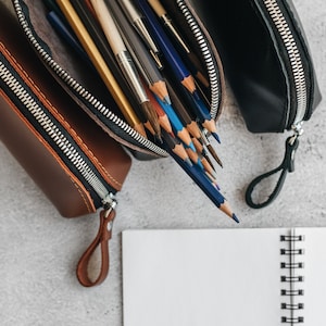 Op de afbeelding: Drie leren etuis, bruin en zwart, met zilveren ritsen en bijpassende treklipjes, zijn open om potloden en penselen te onthullen. Een wit spiraalgebonden notitieboek ligt op de voorgrond op een grijze ondergrond.