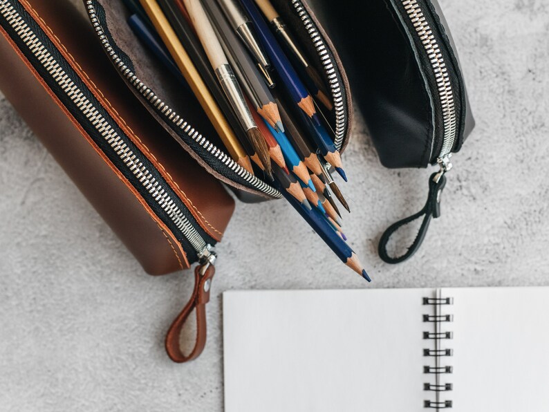 Op de afbeelding: Drie leren etuis, bruin en zwart, met zilveren ritsen en bijpassende treklipjes, zijn open om potloden en penselen te onthullen. Een wit spiraalgebonden notitieboek ligt op de voorgrond op een grijze ondergrond.