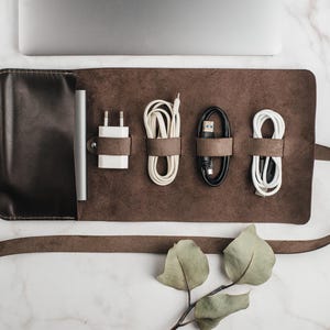 May include: A brown leather roll-up case for organizing and storing cords and chargers. The case has a flap that closes with a snap and a strap that wraps around the case. The case is empty and has five slots for cords and chargers.