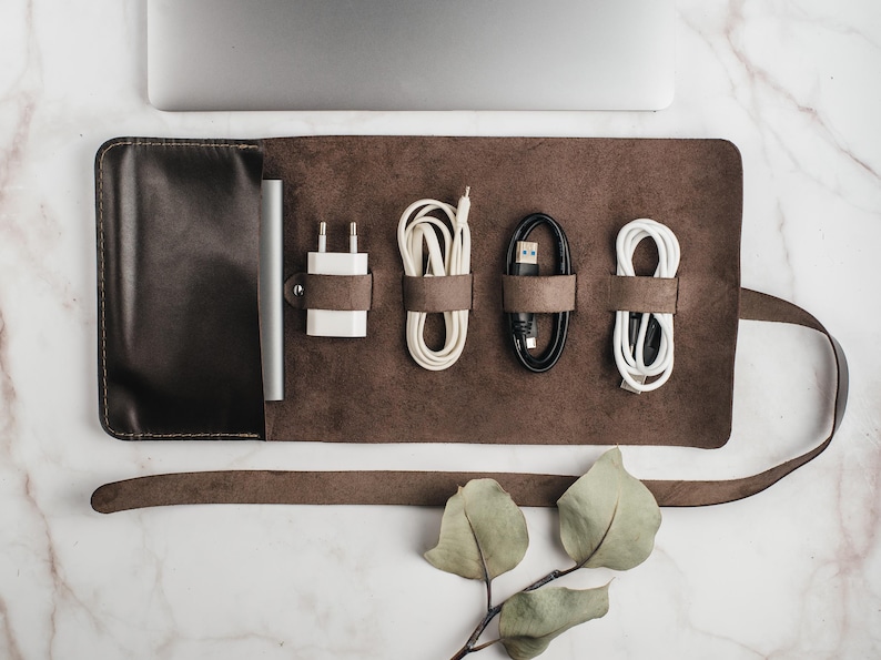 May include: A brown leather roll-up case for organizing and storing cords and chargers. The case has a flap that closes with a snap and a strap that wraps around the case. The case is empty and has five slots for cords and chargers.