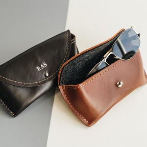 Puede incluir: Dos estuches de cuero para gafas con cierre de broche. Un estuche es de color marrón oscuro con las letras RAS grabadas en la solapa frontal. El otro estuche es de color tostado claro y contiene unas gafas de sol con montura plateada.