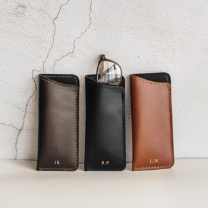 Puede incluir: Tres fundas de cuero para gafas en marrón oscuro, negro y tostado, con costuras visibles en los bordes e iniciales JK, R.P. y E.W. grabadas en la parte inferior. Un par de gafas se encuentra dentro de la funda negra.