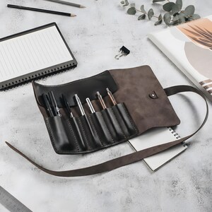 May include: A dark brown leather roll-up pen case featuring six individual slots containing various pens. The case includes a long leather strap for securing the roll and a metal snap button closure. The interior is lined with a smooth, dark material.
