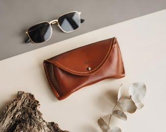 May include: A cognac-colored leather glasses case with a curved flap, a silver-toned metal snap closure, and the embossed letters AR. The case features visible stitching along the edges and is accompanied by gold-rimmed sunglasses.