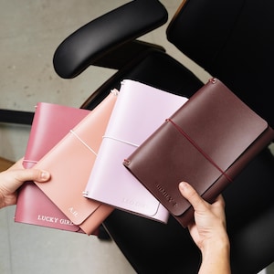 May include: Four rectangular notebooks with smooth covers in shades of dark pink, peach, light purple, and dark brown. Each notebook features a thin elastic band closure and custom text embossed on the front cover, including LUCKY GIRL, A.H., LEO GIRL, and SHMILY.