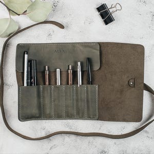 May include: A grey leather roll-up case with a flap closure and a personalised name "ALEX" embossed on the front. The case is open and contains a variety of writing instruments, including pens and pencils.