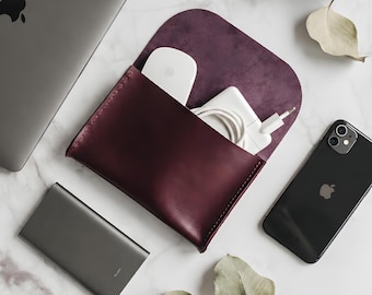 May include: A deep burgundy leather pouch with a flap closure, containing a white computer mouse and a white power adapter with a coiled cable. The pouch is surrounded by a gray laptop, a black smartphone, a gray power bank, and dried leaves.