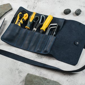 Puede incluir: Un estuche de herramientas de cuero azul oscuro con múltiples bolsillos cosidos que contiene un cúter, un destornillador con mango amarillo, unas tijeras, otro destornillador amarillo y dos llaves hexagonales de metal.