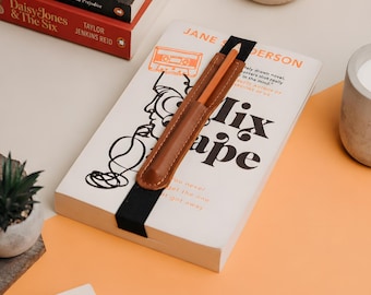 May include: A brown leather pen holder attached to a black elastic band, secured around a white paperback book titled Mixtape by Jane Sanderson. The holder contains a single orange pencil, positioned vertically against the book cover.