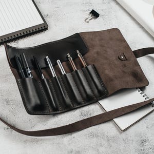 May include: A brown leather pen roll with a strap closure. The roll has eight slots for pens and pencils. The roll is lying on a white surface with a notebook and a paperclip.