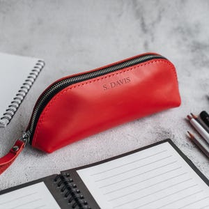 Op de afbeelding: Een rode leren etui met een zwarte ritssluiting en een polsbandje. De tekst "S. DAVIS" is in reli&euml;f op de voorkant aangebracht. De etui ligt naast een spiraalgebonden notitieboek, potloden en penselen.