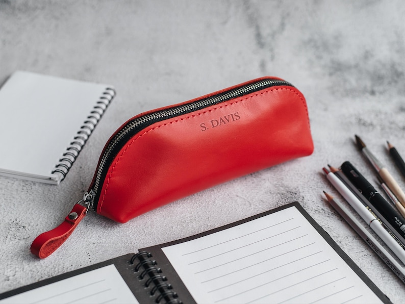 Op de afbeelding: Een rode leren etui met een zwarte ritssluiting en een polsbandje. De tekst "S. DAVIS" is in reli&euml;f op de voorkant aangebracht. De etui ligt naast een spiraalgebonden notitieboek, potloden en penselen.