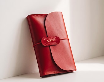 May include: A rectangular red leather journal cover with a curved flap closure. A thin red elastic band wraps around the center, securing the flap with a small leather tab embossed with the gold-colored capital letters A.R.J.