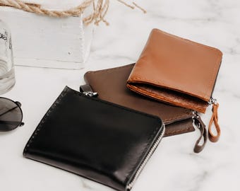 May include: Three rectangular wallets with zipper closures, arranged in a stack. The wallets are made of smooth material in black, dark brown, and tan colors. Each wallet features a matching pull tab attached to the silver-toned metal zipper.