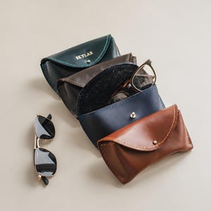 Puede incluir: Una colección de cuatro estuches de cuero para gafas en verde oscuro, marrón oscuro, azul marino y color canela, cada uno con cierre de broche metálico y costuras visibles. El estuche verde tiene grabado el nombre SKYLAR. Unas gafas están al lado.