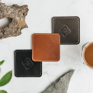 Può includere: Tre sottobicchieri quadrati in pelle nera, marrone e beige, con bordi cuciti. Ogni sottobicchiere presenta un emblema a forma di diamante con testo. Un bicchiere di caffè è visibile sullo sfondo.