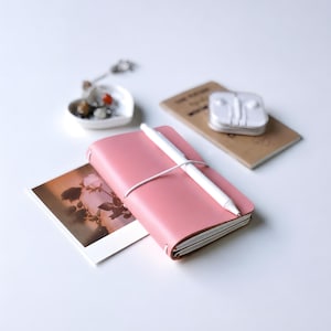 May include: A pink leather-bound notebook with a white elastic band closure and a white pen resting on top. A rectangular photograph of roses sits beneath the notebook, while a small heart-shaped dish with charms and white earbuds are nearby.