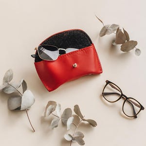 May include: A bright red leather glasses case with a silver snap closure, containing a pair of silver-framed sunglasses. A second pair of black-rimmed glasses rests nearby, accompanied by several sprigs of dried eucalyptus leaves.