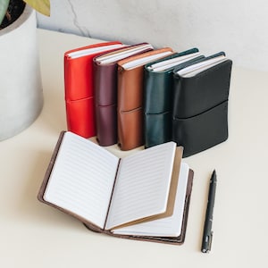 Puede incluir: Varios cuadernos de cuero en rojo, morado, marrón, verde y negro. Uno está abierto, mostrando páginas rayadas. Un bolígrafo negro está al lado, ideal para tomar notas o escribir un diario.