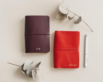 May include: Two leather-bound journals with elastic closures. The journal on the left is burgundy with the initials "E.W." embossed in white. The journal on the right is red with the initials "SMCJH" embossed in white.