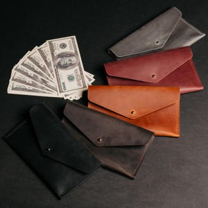 May include: An assortment of leather envelope wallets in various colors, including black, brown, and gray, with a fan of hundred-dollar bills. Each wallet features a snap closure. The wallets are arranged on a dark surface.