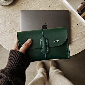 May include: A dark green leather pouch with a rounded flap closure secured by a thin elastic cord and a matching leather tab. The front of the pouch features the embossed white letters MJW and visible green stitching along the edges.