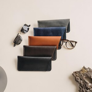 May include: A collection of five rectangular leather eyeglass cases in gray, blue, tan, dark brown, and black, each featuring visible stitching along the edges. Two pairs of eyeglasses are positioned near the cases, one with gold frames.