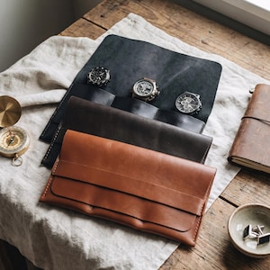 May include: Three rectangular leather watch rolls in black, dark brown, and tan, each featuring a flap closure and stitched edges. The black roll is open, displaying three wristwatches with round faces and metallic details arranged in a row.