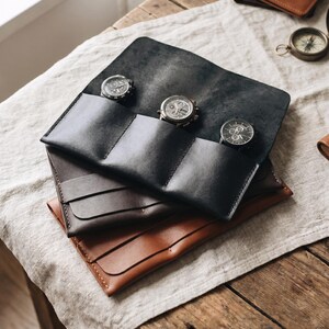 Puede incluir: Un estuche enrollable de cuero negro con tres compartimentos, cada uno con un reloj plateado. El estuche está abierto, revelando los relojes, y descansa sobre otros dos estuches enrollables de cuero marrón y gris. Los artículos están sobre una superficie de madera.