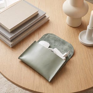 May include: A sage green leather pouch with a flap closure, containing a white computer mouse and a white charging cable with a power adapter. The pouch features smooth, stitched edges and a soft, matte finish on the interior flap surface.