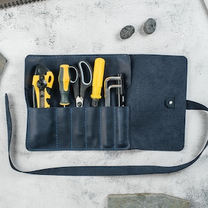 May include: A navy blue leather tool roll, open to display various hand tools. The tools include a yellow and black wire stripper, screwdrivers, scissors, and Allen wrenches. The roll has a strap for carrying.