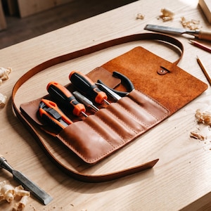 Op de afbeelding: Een bruine lederen gereedschapsetui met meerdere vakken waarin een stanleymes, twee schroevendraaiers met oranje en zwarte handgrepen en een tang zitten. De etui heeft een lange leren riem en een metalen drukknoopsluiting.