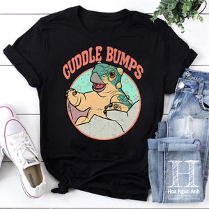 Puede incluir: Camiseta negra con un diseño de dinosaurio de dibujos animados y el texto "Cuddle Bumps". El dinosaurio es de color marrón claro y verde azulado, dentro de un círculo azul claro con estrellas. Un diseño divertido y casual.