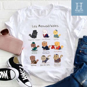 Puede incluir: Camiseta blanca con el texto "Les Meowsérables" e ilustraciones de gatos disfrazados. Cada gato tiene un nombre debajo. También se ven unas zapatillas negras y blancas y una cartera rosa.