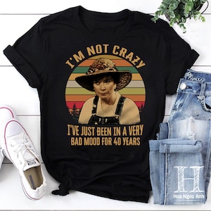 May include: A black t-shirt with a vintage-style graphic of a woman with a straw hat. The graphic includes the text "I'm not crazy I've just been in a very bad mood for 40 years".