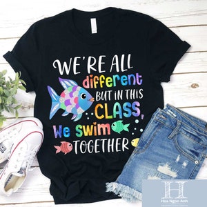 Puede incluir: Camiseta negra con un gráfico de pez colorido y el texto "We're all different but in this class, we swim together".