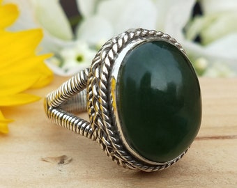 May include: A silver ring with a large, oval, dark green gemstone. The ring has a braided design around the gemstone and on the band. The ring is displayed on a wooden surface with a yellow flower and white flowers in the background.