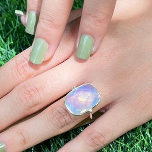 Puede incluir: Un anillo de plata con una gran piedra ovalada iridiscente. El anillo est&aacute; en una mano con esmalte de u&ntilde;as verde.