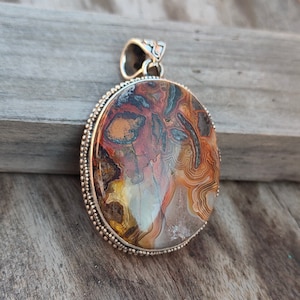 May include: A silver pendant featuring a round, brown, white and red agate stone. The stone has a swirling pattern and is set in a silver bezel with a beaded edge.