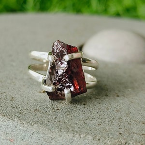 May include: A silver ring with a raw, red garnet gemstone. The ring has a simple, modern design with a double band.
